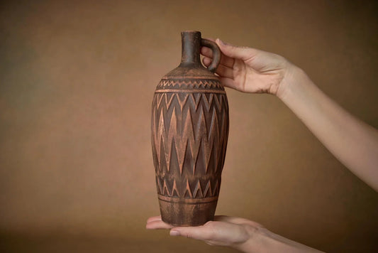 Handmade brown clay vase with carved geometric patterns held by two hands
