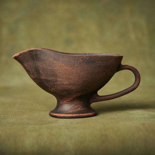 Handmade brown clay gravy boat with a rustic textured finish on a green fabric background.