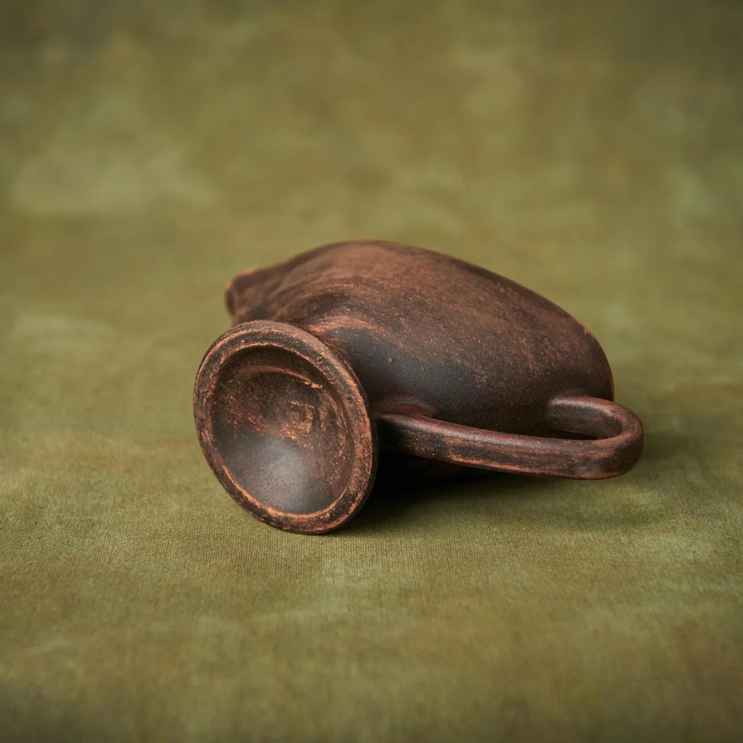 Handmade brown clay pitcher with a rustic texture on a green fabric background.