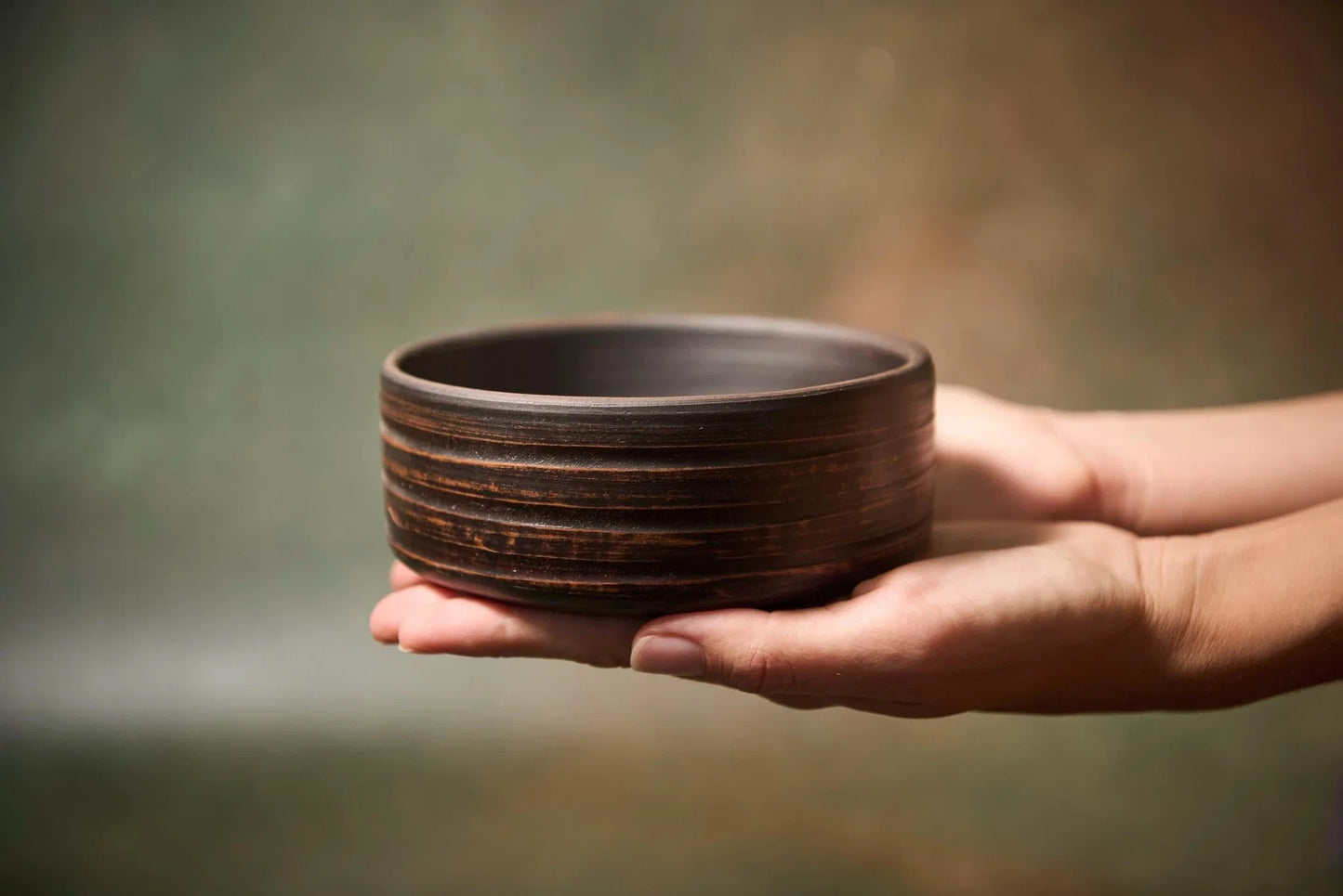 Hand holding a handmade dark brown clay bowl with horizontal textured ridges