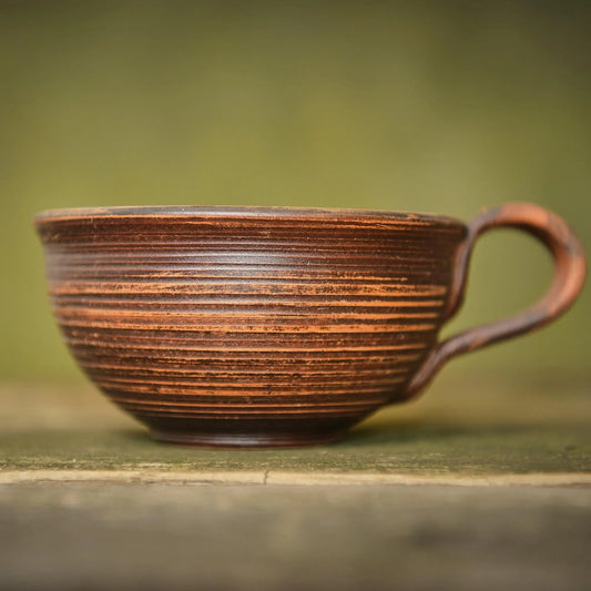 Handmade brown clay mug with textured horizontal ridges on a rustic wooden surface