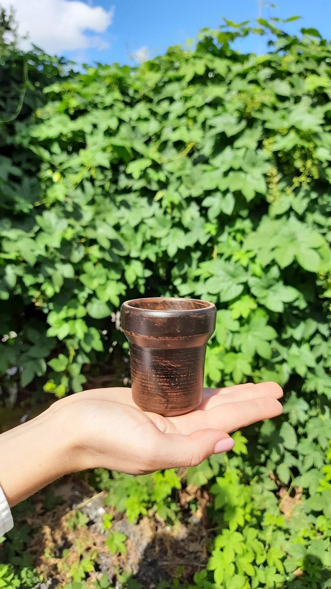 Handmade dark brown clay cup held in a hand against a lush green garden background.