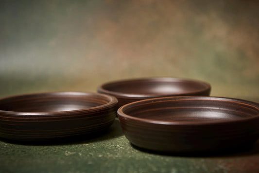 Three handmade brown clay shallow bowls on a textured green background