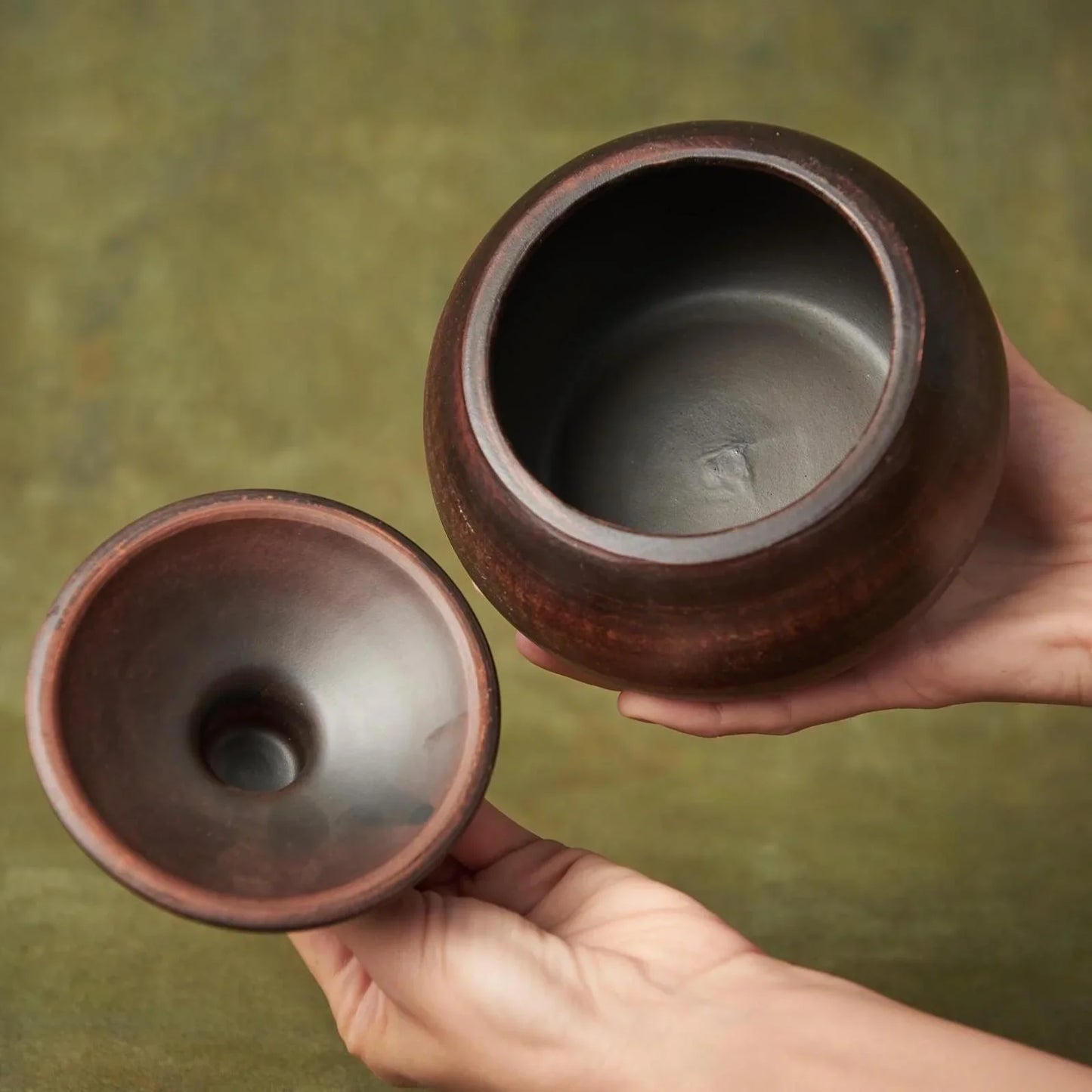 Hands holding two handmade dark brown clay pottery vessels against a green background.
