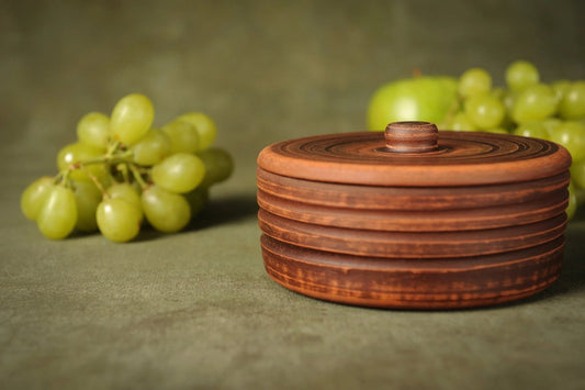 Handmade rustic clay pot with lid on green background, grapes and apple, clayproductsshop homeware