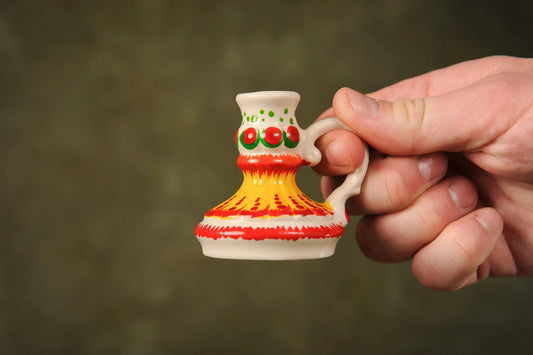 Hand holding a handmade, hand-painted ceramic candlestick with colorful red, yellow, and green patterns, artisanal white clay candelabrum from clayproductsshop.