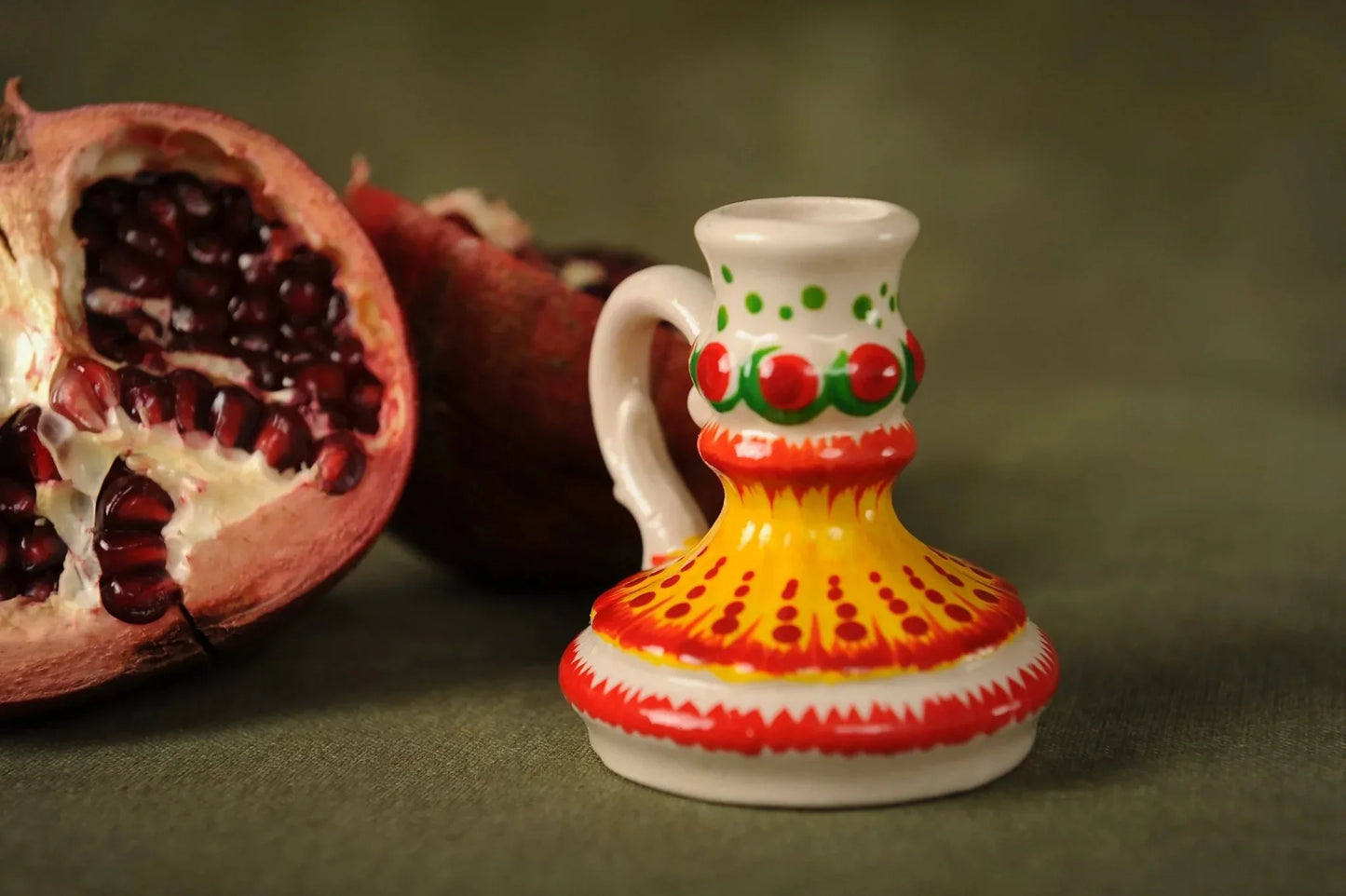 Hand-painted ceramic candlestick in vibrant yellow, red, and green sits beside a pomegranate, artisan home decor from clayproductsshop.
