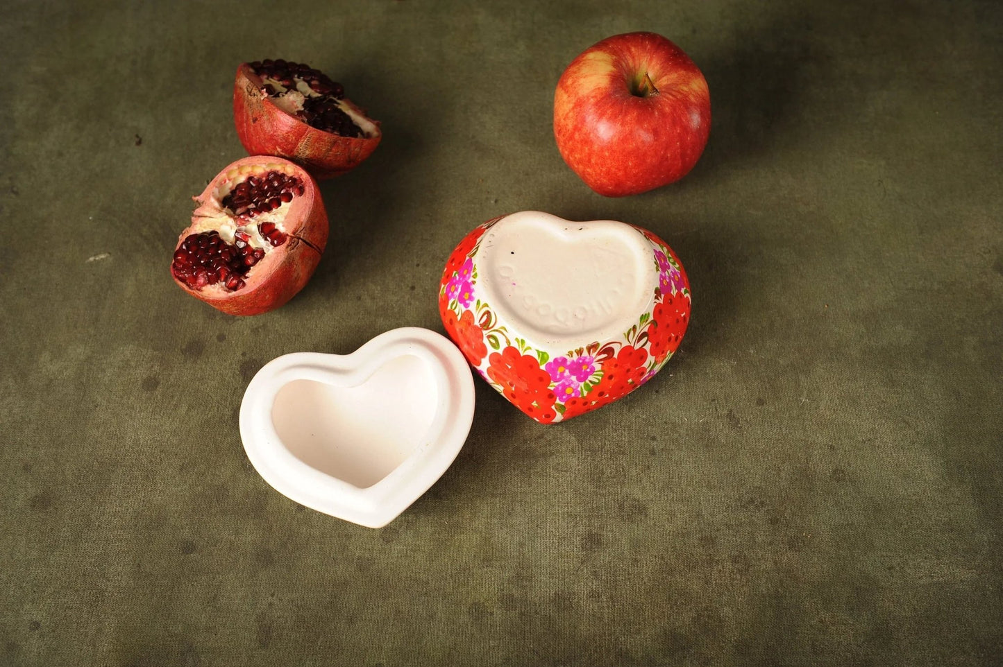Hand-painted heart-shaped ceramic box with floral design, open on a table with an apple and pomegranate