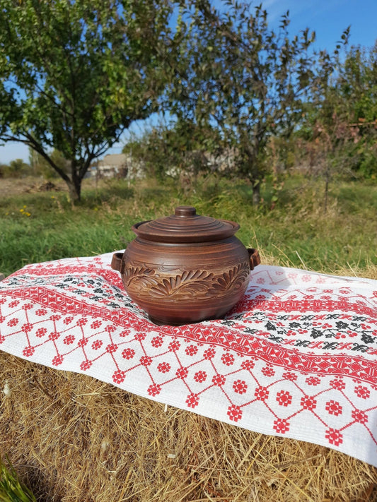 Handmade ceramic pot with lid and handles on embroidered cloth outdoors, clay kitchenware