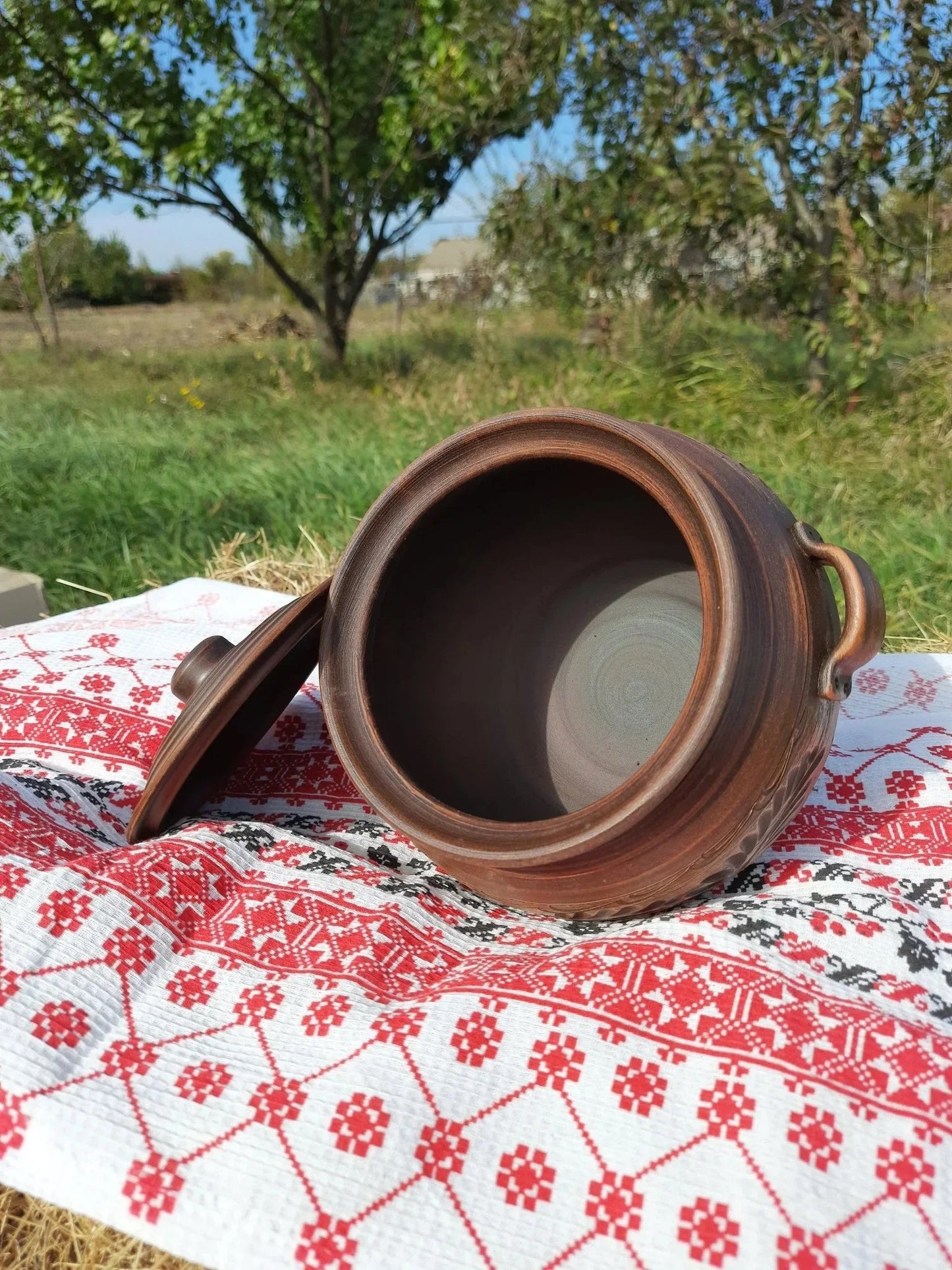 Handmade red clay ceramic pot with lid and handles on embroidered cloth outdoors