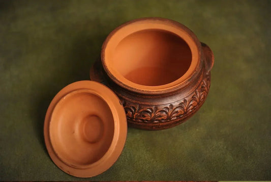 Handmade brown clay pot with ornate pattern and lid on green background, artisan homeware