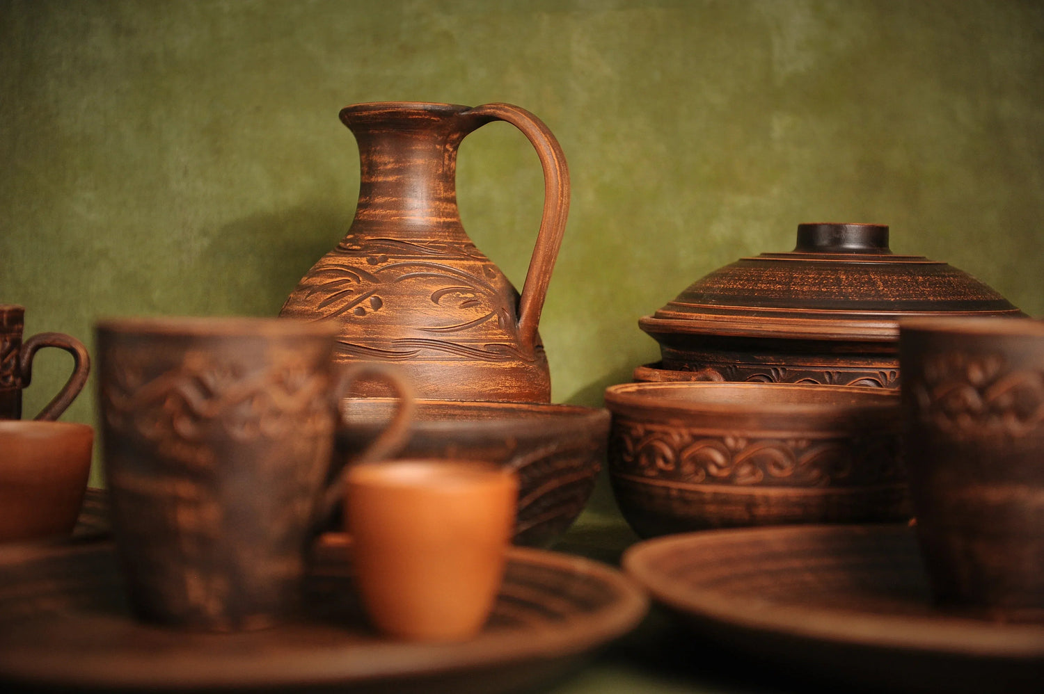 Handmade clay pottery set with cups, plates, jug, and bowls on green background, artisan homeware