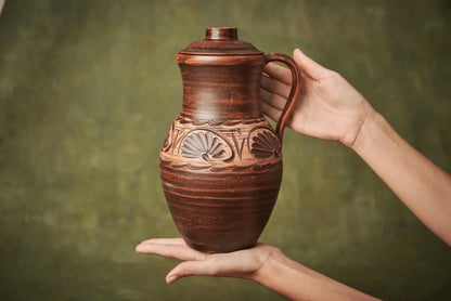 Hand holding handmade clay jug with lid, artisan ceramic vessel with carved patterns