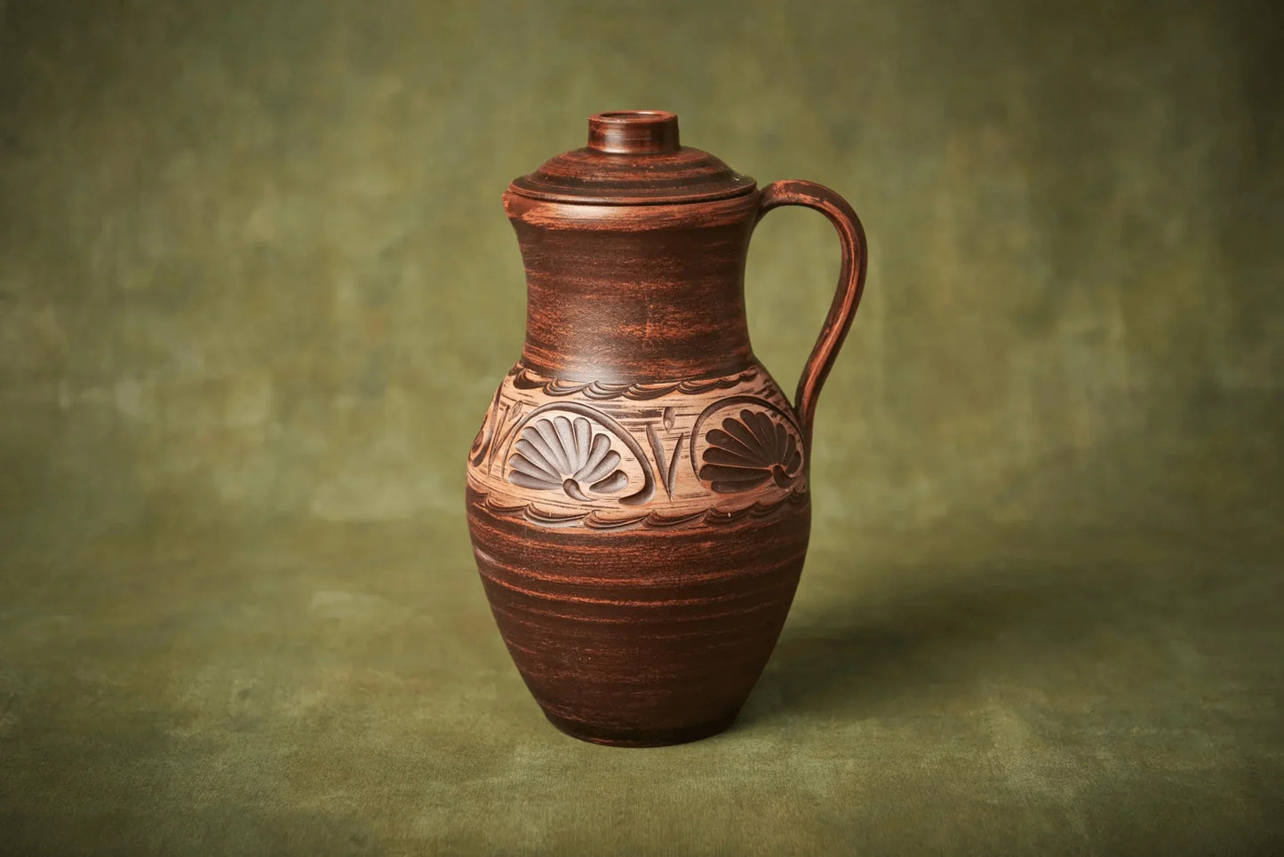 Handmade clay jug with lid and carved design, artisan ceramic homeware on green background