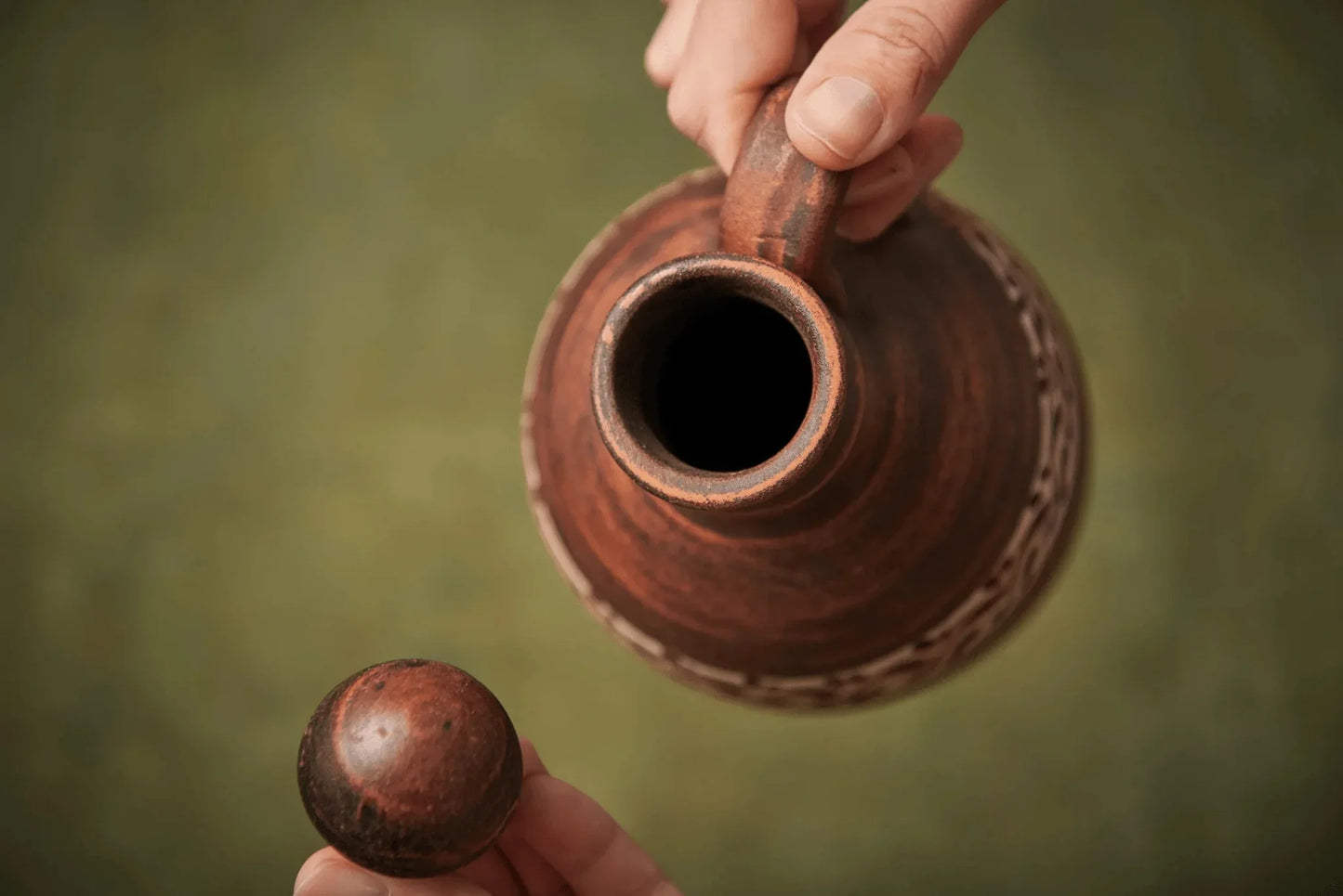 Hand lifting the lid from a brown handmade ceramic clay amphora jug with handle