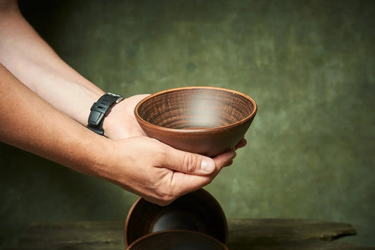 Handmade deep red clay ceramic bowl held in hands, rustic Ukrainian pottery on wood table