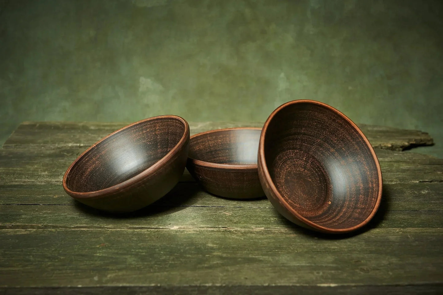 Set of three handmade deep red clay ceramic bowls on rustic wooden table, artisan pottery