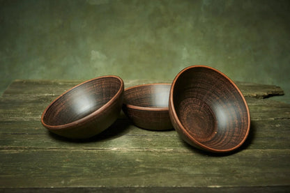 Set of three handmade deep red clay ceramic bowls on rustic wooden table, artisan pottery