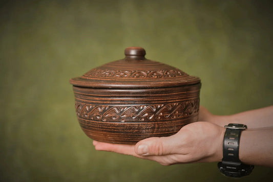 Hand holding a carved handmade ceramic clay bread pot with lid against a green background