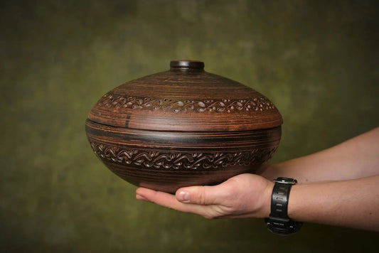 Hand holding a round handmade clay cooking pot with lid and decorative carvings
