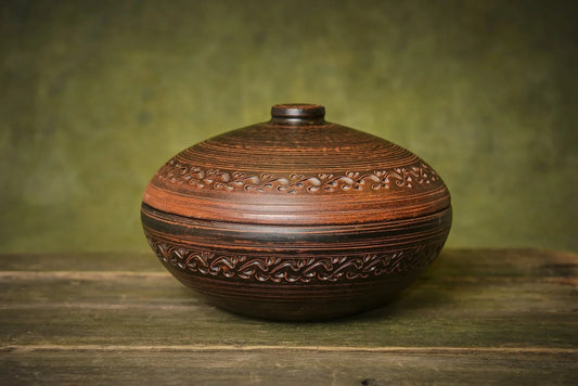 Handmade round clay cooking pot with lid and decorative carvings on rustic wood table