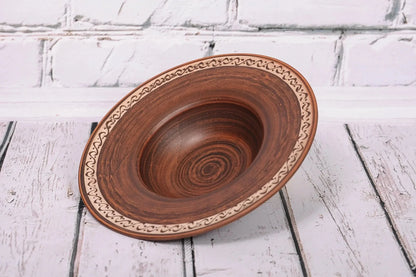 Handmade ceramic pasta bowl with decorative rim, rustic brown clay, on white wooden surface