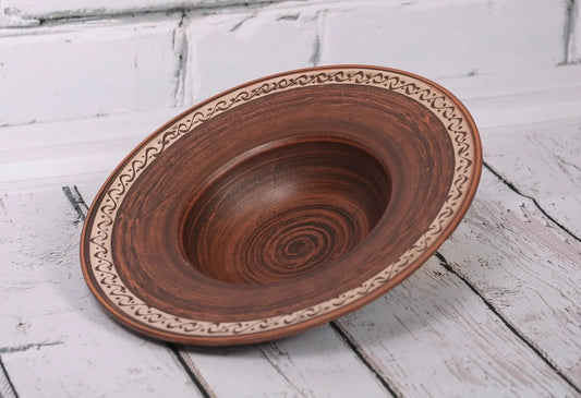 Handmade brown ceramic pasta bowl with carved border on white wooden surface