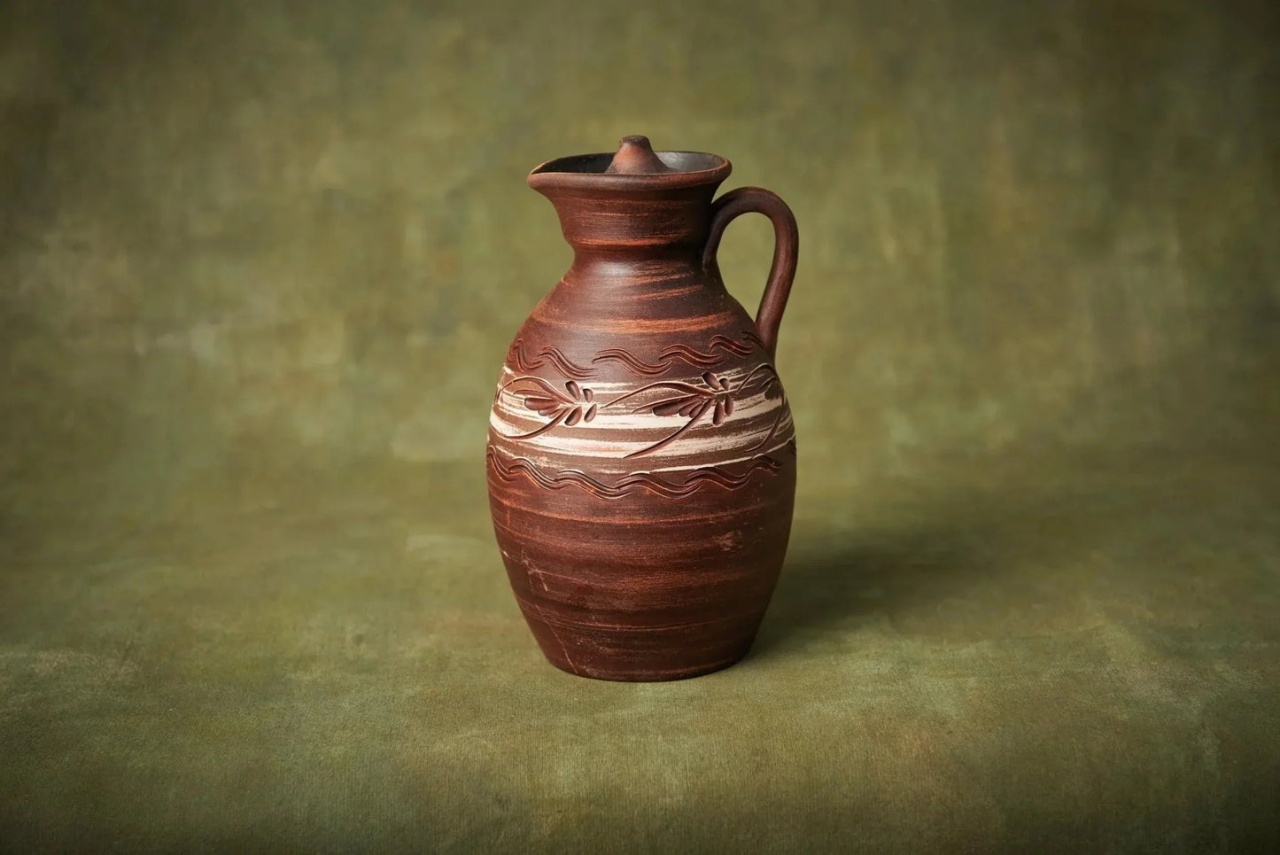 Handmade clay ceramic pitcher with lid and floral engraving on green background