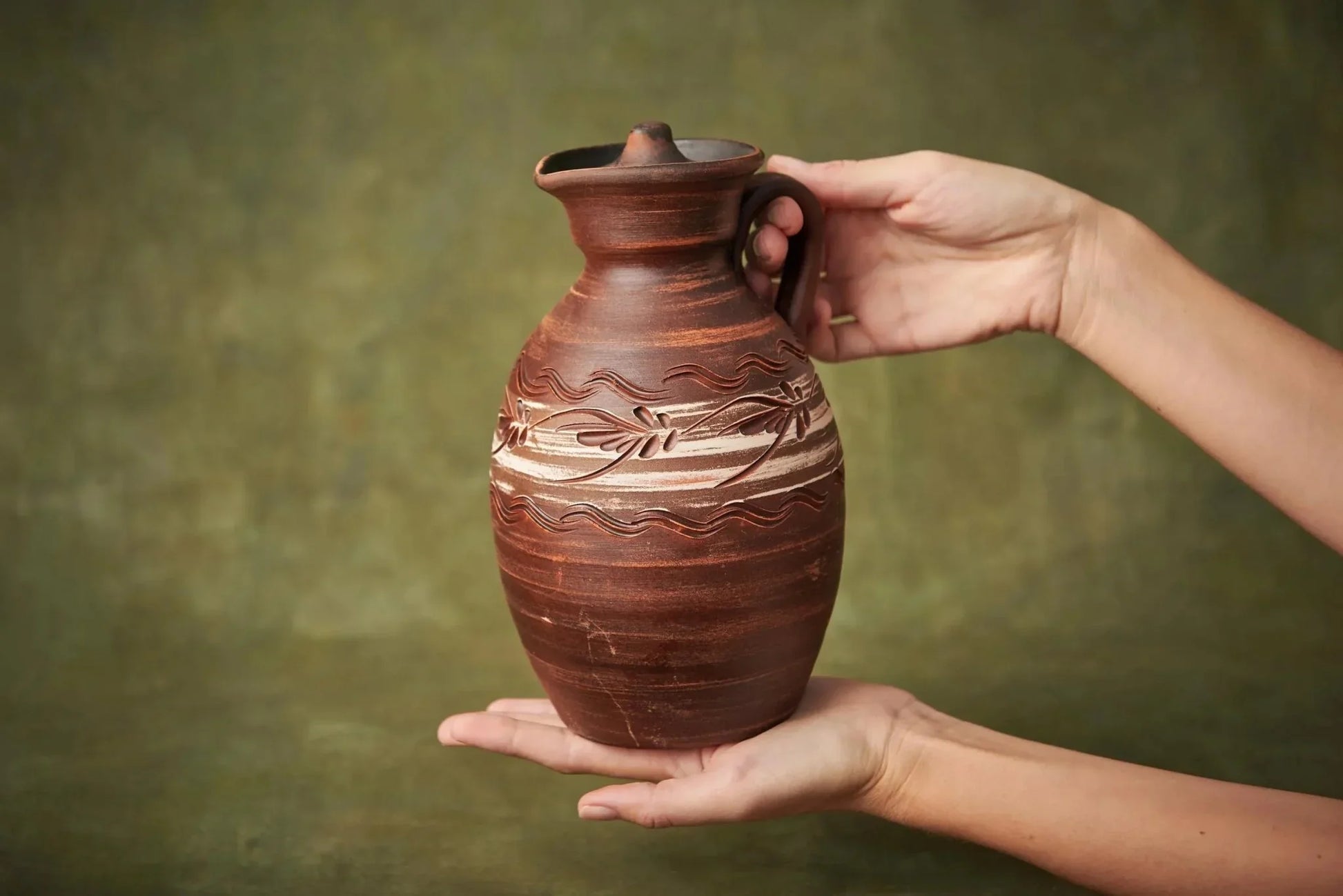 Handmade clay ceramic pitcher with lid and decorative carved pattern, held in hands