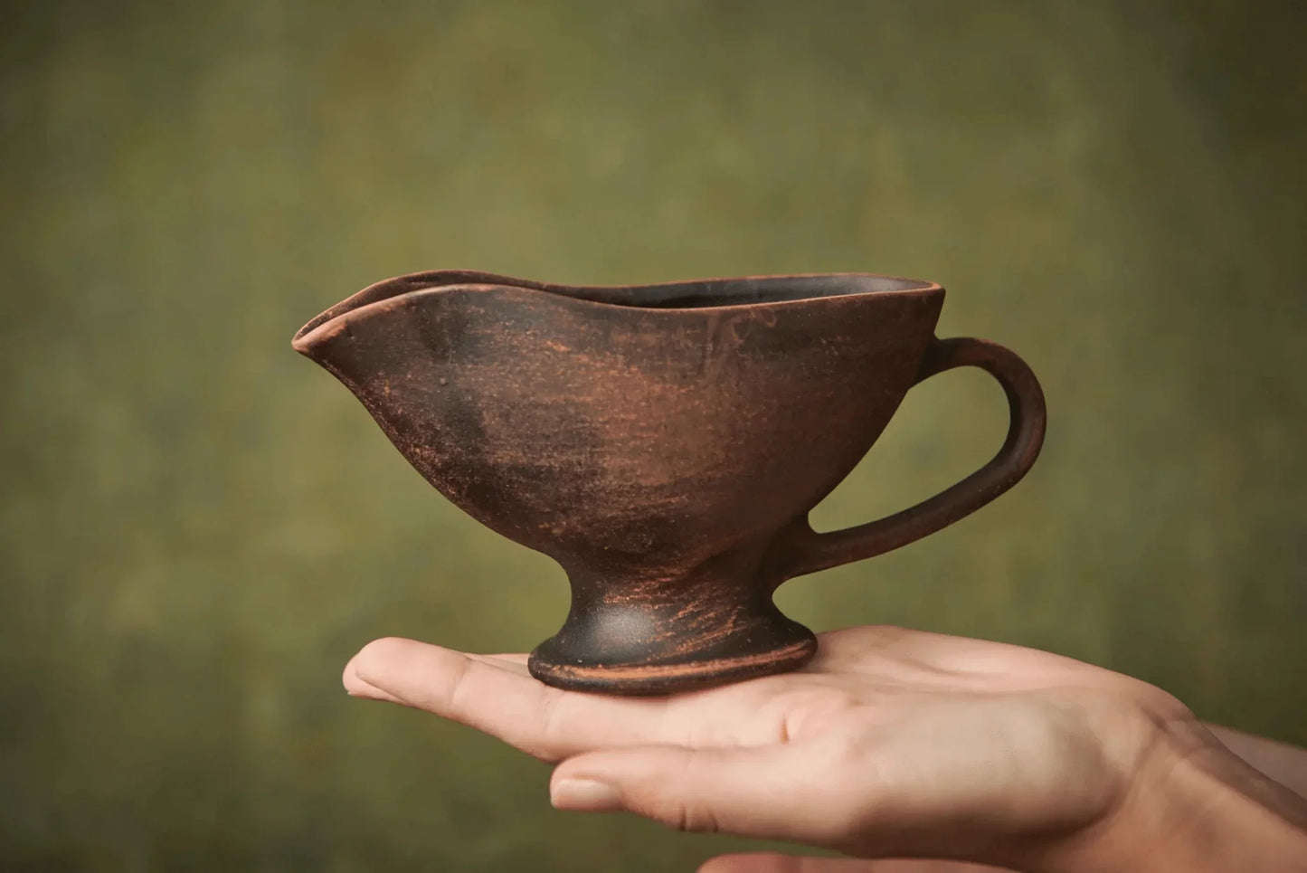 Handmade ceramic sauceboat in brown clay, held in a hand against a green background