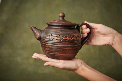 Hand holding a handcrafted brown ceramic teapot with engraved clay design, artisan home decor