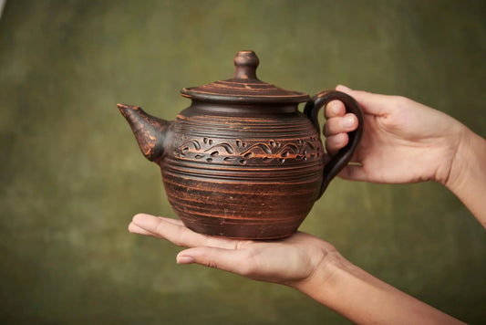 Hand holding a handcrafted brown ceramic teapot with engraved clay design, artisan home decor