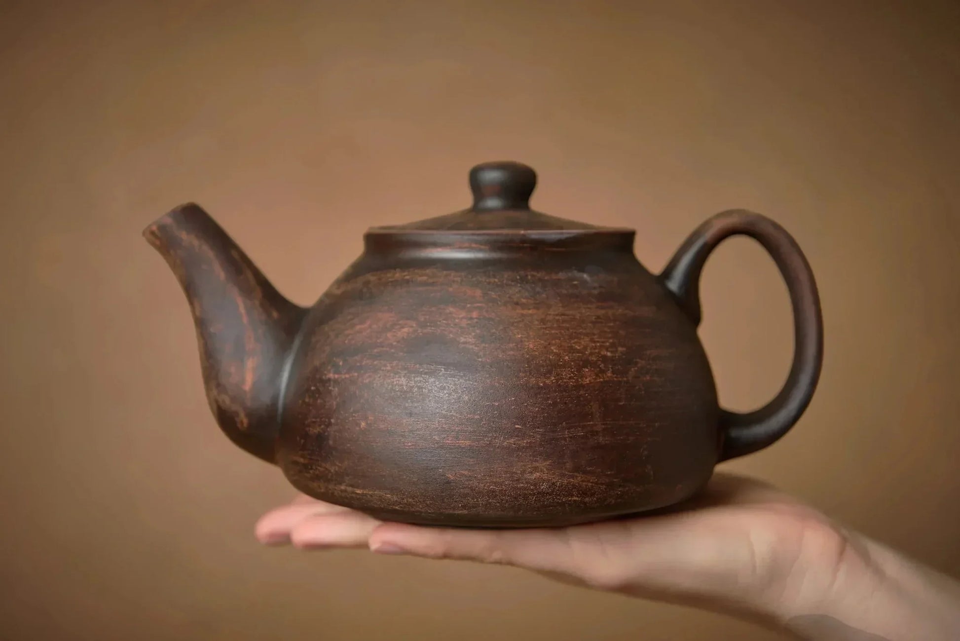 Handmade brown ceramic teapot with lid, artisan pottery, held in a hand against neutral background