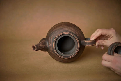 Hand holding artisan handmade ceramic teapot, rustic brown clay pottery, on neutral background