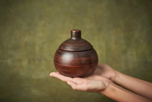 Hand holding a small handmade ceramic clay baking pot with lid against a neutral background