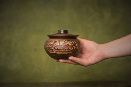 Hand holding a handmade ceramic sugar bowl with lid and floral carving, rustic clay pottery.