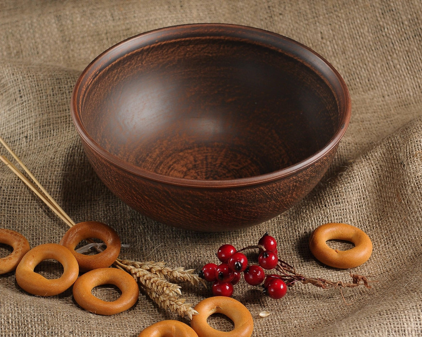 Handmade brown clay bowl with rustic finish, surrounded by bagels and red berries on burlap