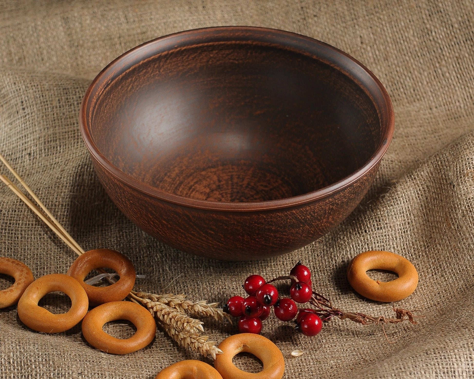 Handmade brown clay bowl with rustic finish, surrounded by bagels and red berries on burlap