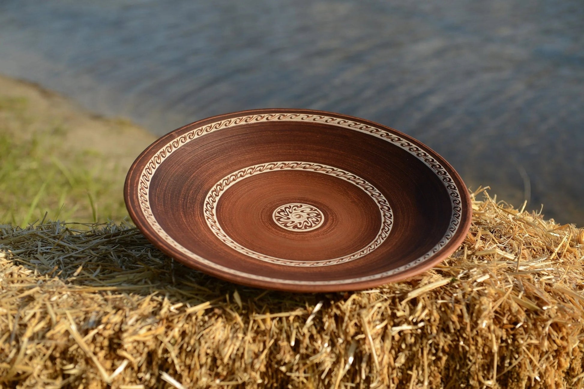 Handmade large brown ceramic plate with decorative rim, displayed outdoors on straw by water