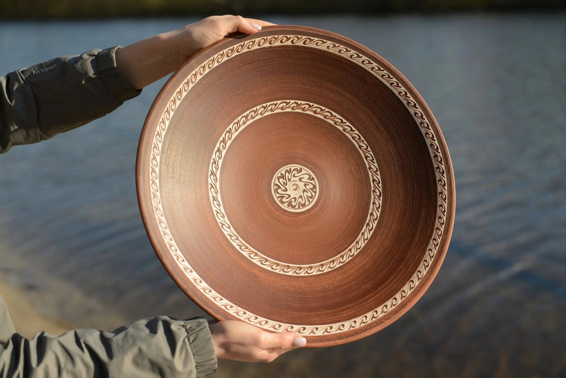 Hand holding large handmade red clay ceramic plate with decorative patterns by water