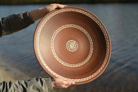 Hand holding large handmade red clay ceramic plate with decorative patterns by water