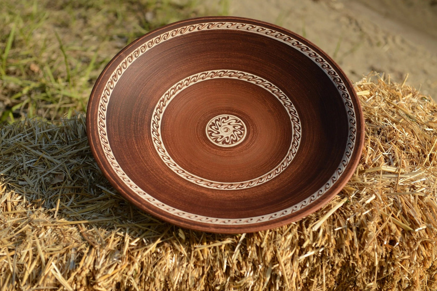 Handmade large brown ceramic clay plate with decorative patterns, outdoor on straw bale
