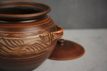 Handmade clay casserole baking dish with carved pattern and lid on gray surface