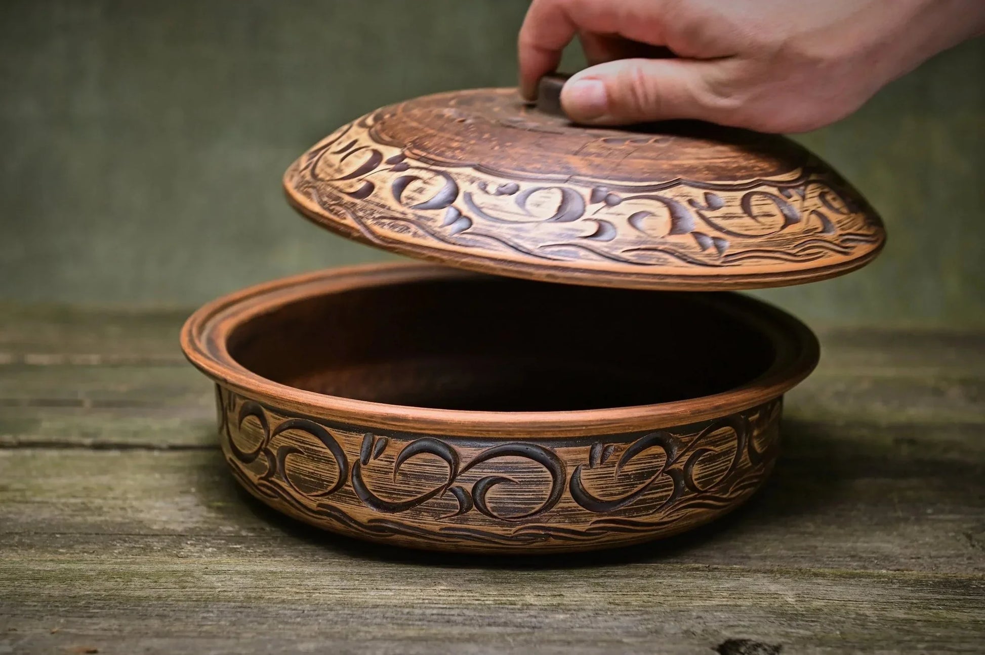 Hand lifting lid of handmade ceramic clay baking pot with decorative carvings on rustic wood