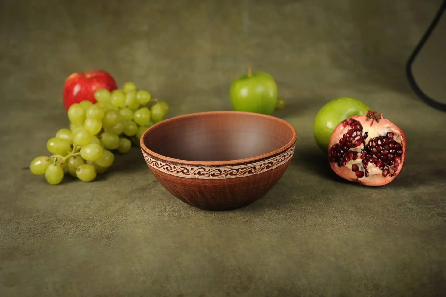 Handmade red clay ceramic bowl with rustic decorative rim, surrounded by fruits on a green surface