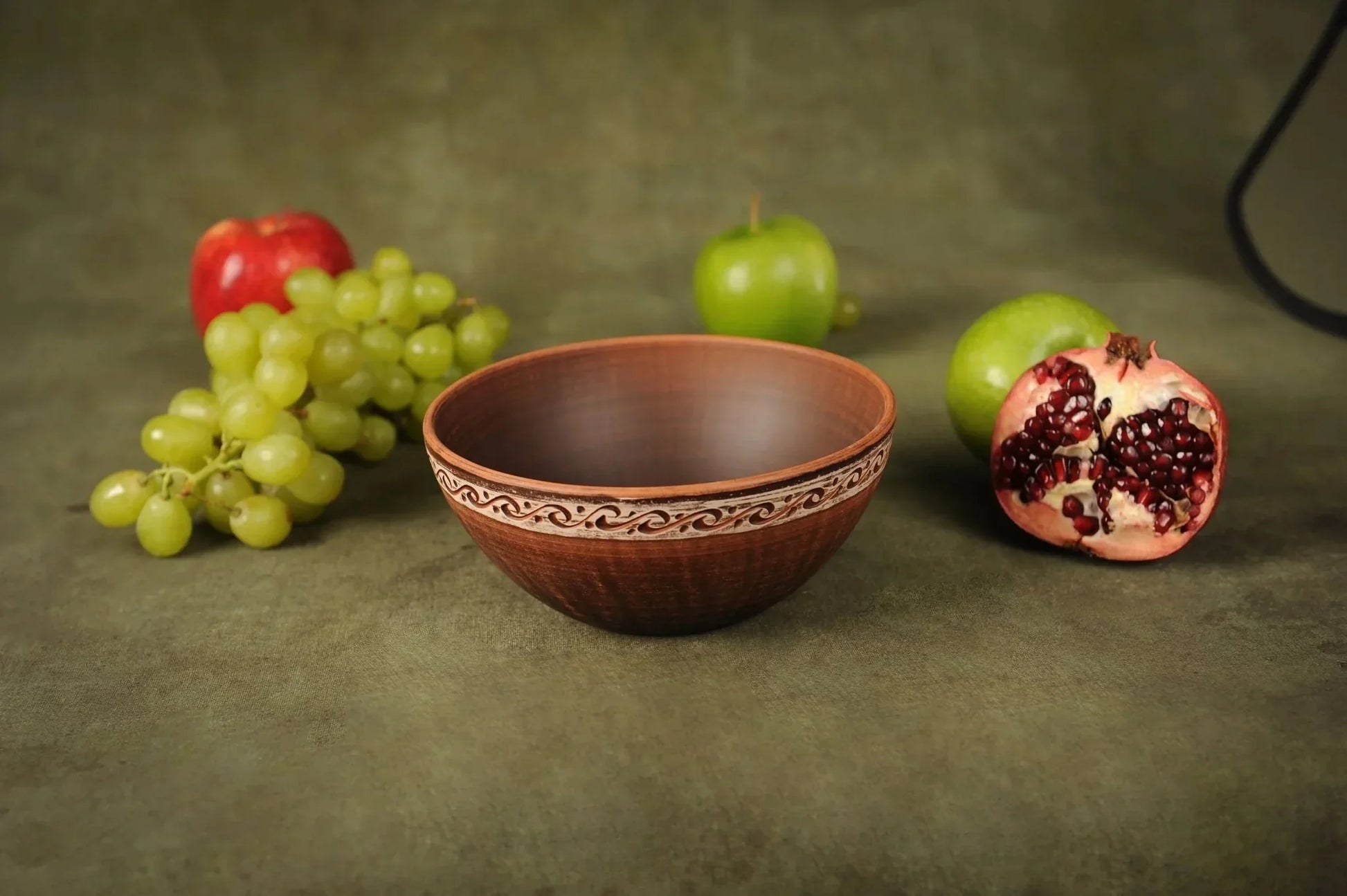 Handmade red clay ceramic bowl with rustic decorative rim, surrounded by fruits on a green surface