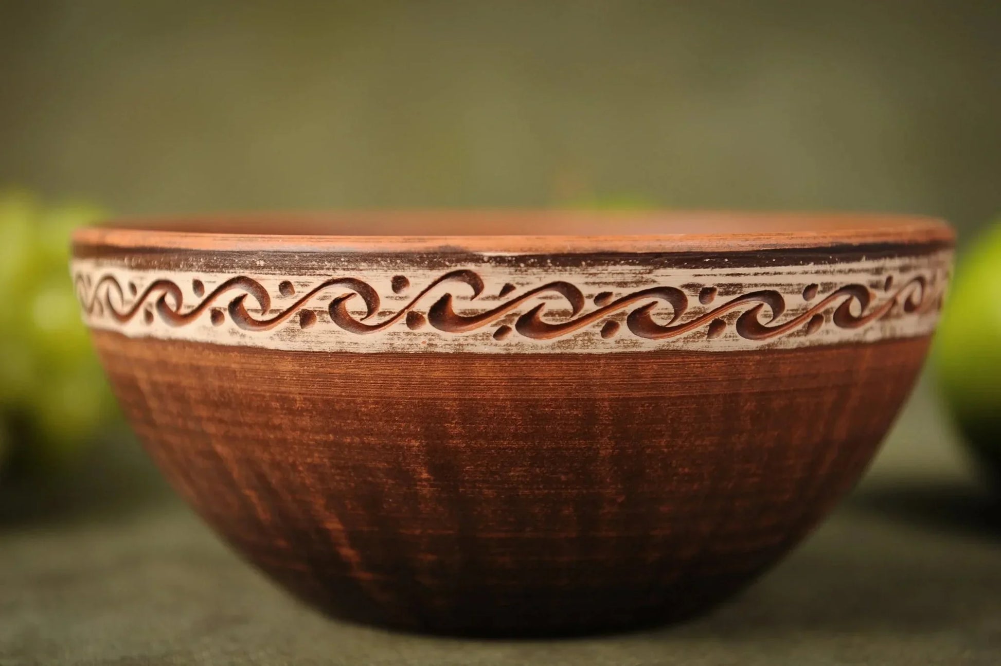 Handmade brown ceramic clay bowl with decorative carved rim on green surface