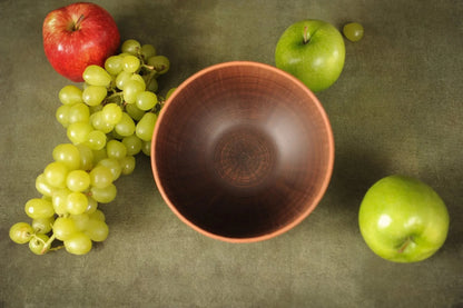 Handmade brown ceramic bowl with apples and green grapes on a rustic surface
