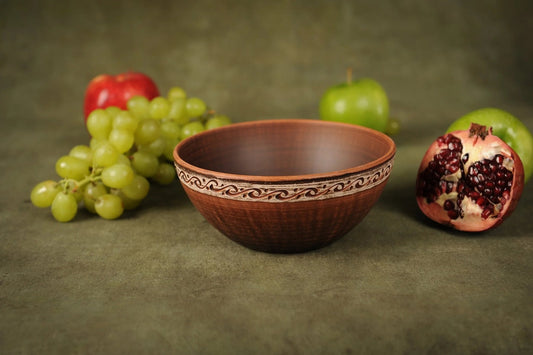 Handmade rustic red clay ceramic bowl with carved rim, displayed with fruit on green background
