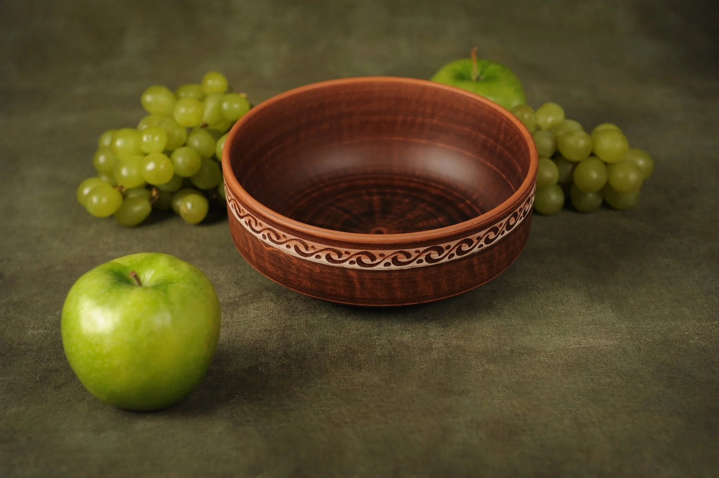 Handmade rustic clay bowl with decorative pattern, surrounded by green apples and grapes.